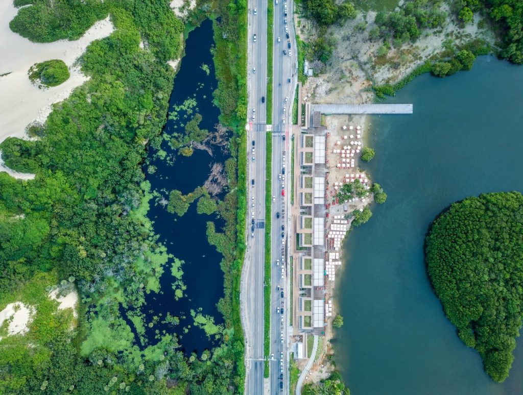 Vista aérea de área urbana litorânea em Fortaleza, com estrada, vegetação e faixa costeira.